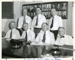 Image of several men wearing ties and white shirts. There are photographs of men in suits behind them, and a wooden table with notepads, books, and empty ashtrays in front of them.