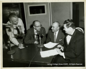 Several men with papers in front of them, seated at a desk, and talking into microphones