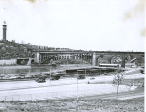 Image of High Bridge, a high way with cars and trucks, a distant wter tower and houses of Washington Heights.
