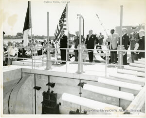 Image of men in suits on a platform wiht American flags. One man is speaking into a microphone.