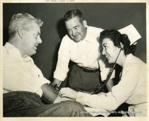 Image of a female nurse checking the blood pressure and arm of a male blood donor. Another man looks on at him, smiling.