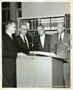 Image of several men in suits looking through bound volumes on a podium. One holds up a book labeled "Poor's Register of Directors and Executives 1958."