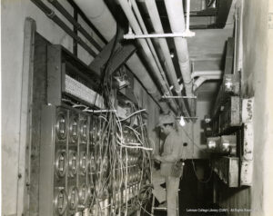 Image of a man kneeling on a step-stool, illuminated by an electric light bulb surrounded by wires, conduits, and electrical infrastructure.