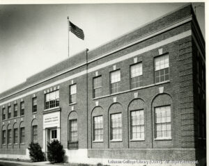 Image of a brick building with an American flag. Two bushes are in front of the door labeled "Manhattan College Engineering Building."