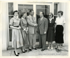 Image of two men in suit shaking hands. Four women in dresses flank them in front of a glass window labeled "New York Telephone Company."