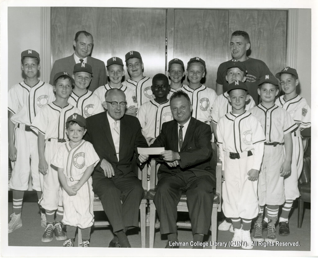 Image of two men in suits passing a check, and several boys in baseball uniforms. One man in a suit is in the back, as is one man in a T-shirt (presumably the coach).