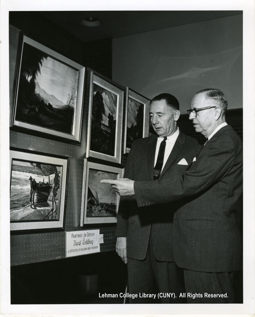 Two men in suits point at paintings on a peg-board.