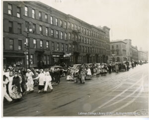 Image of a parade of costumed children. Some are looking at the camera and some are not. They are walking past mixed-use buildings.
