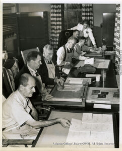 Men in smocks and ties work with silk screen equipment. A women stands over one man, and open curtains are visible.