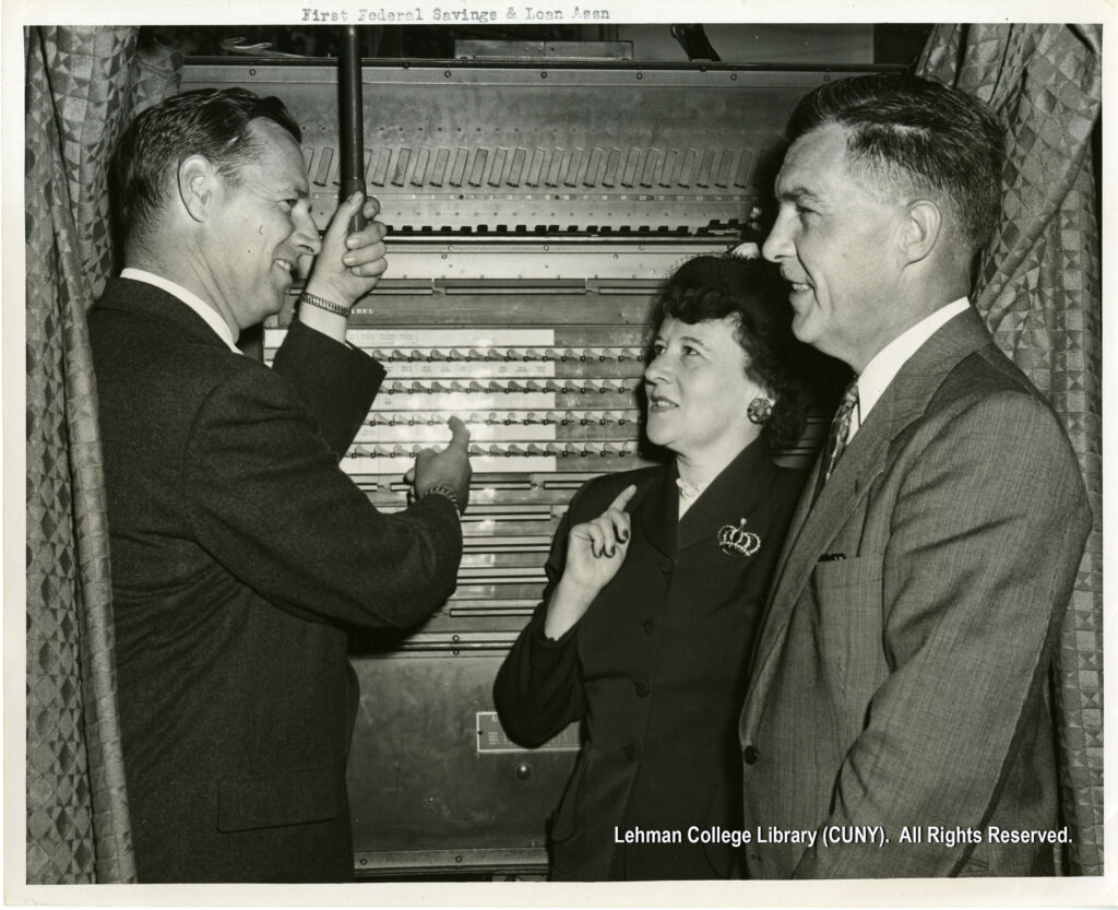 Image of a man in a suit, pointing at a piece of a machine to a man and a woman in suits.