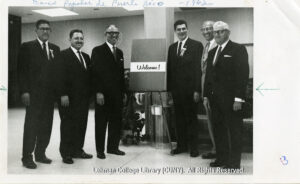 Several men in suits are standing in front of an easel that says "Welcome"